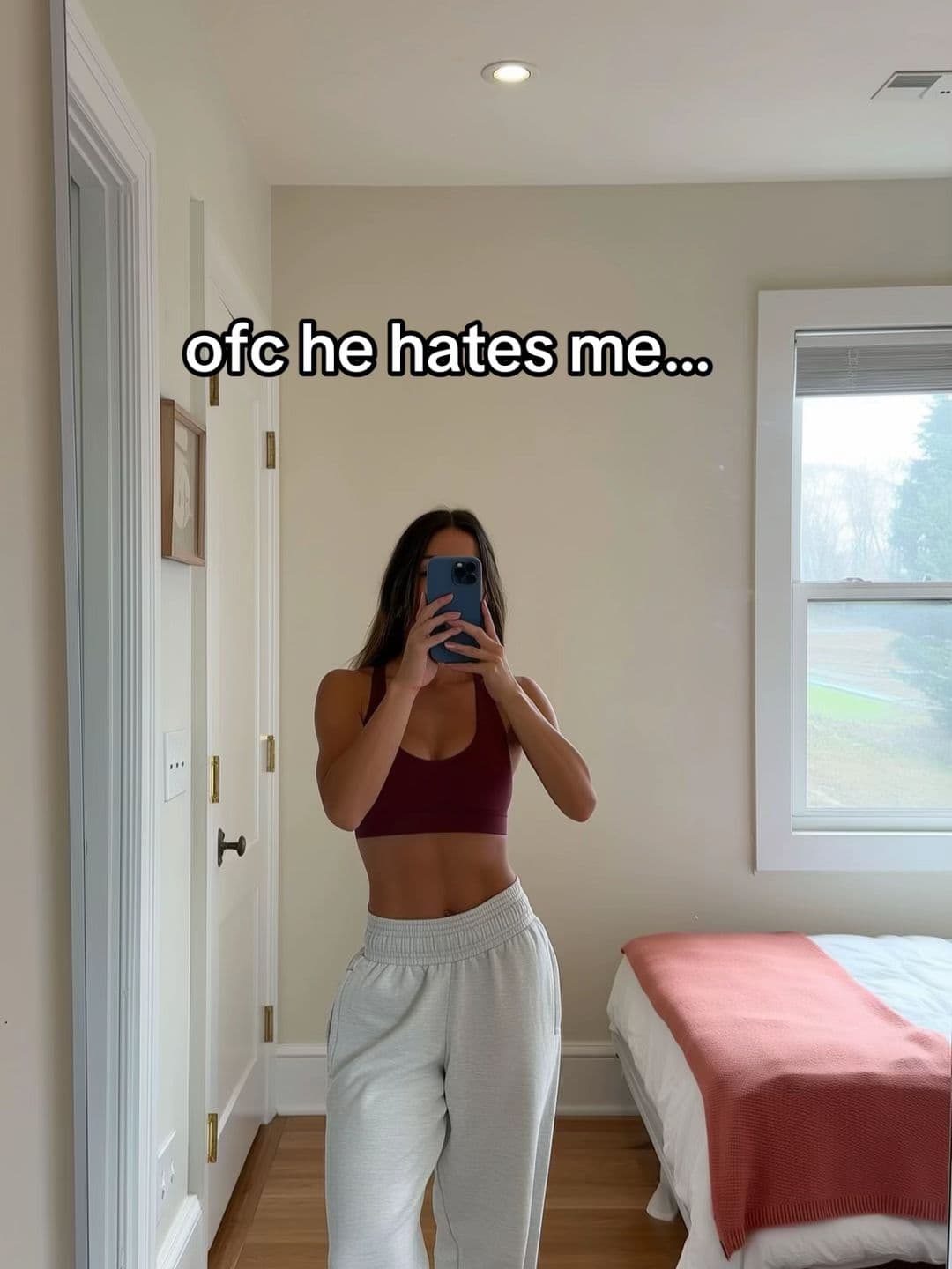 Mirror photo of a woman in gym clothes in a bedroom, with the text ofc he hates me.